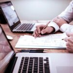 Duas pessoas trabalhando em frente a laptops, analisando documentos e fazendo anotações em uma mesa de escritório.