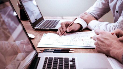 Duas pessoas trabalhando em frente a laptops, analisando documentos e fazendo anotações em uma mesa de escritório.