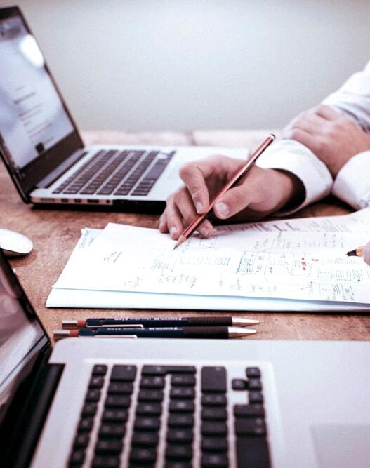 Duas pessoas trabalhando em frente a laptops, analisando documentos e fazendo anotações em uma mesa de escritório.