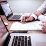 Duas pessoas trabalhando em frente a laptops, analisando documentos e fazendo anotações em uma mesa de escritório.