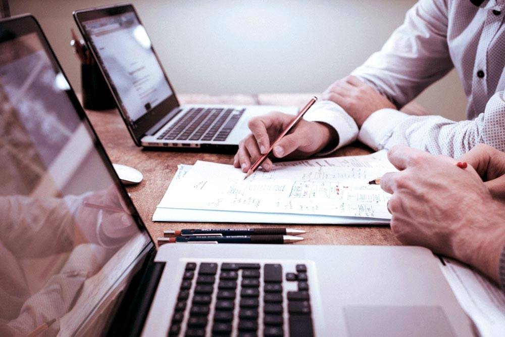 Duas pessoas trabalhando em frente a laptops, analisando documentos e fazendo anotações em uma mesa de escritório.